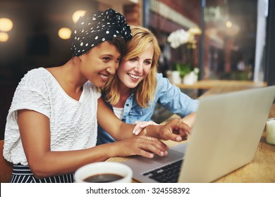 Two multiracial young female friends surfing the internet together on a laptop as they sit in a cafeteria enjoying a cup of coffee - Powered by Shutterstock