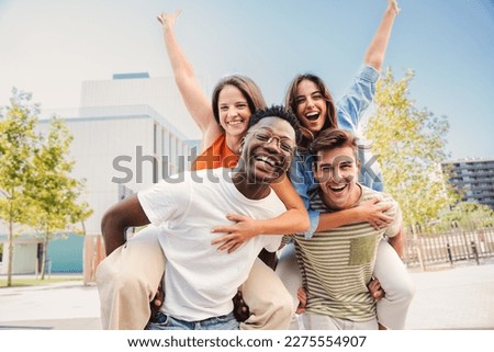 Two multiracial young friends giving piggybacks ride to his girlfriends. A group of happy people having fun and smiling at weekend activity. Men carrying women on his shoulders looking at camera. High