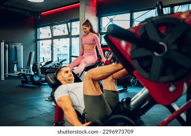 Two muscular gym mates are doing leg exercises on the leg press machines in a gym in different positions. Selective focus on a sportswoman. Sporty couple working out in a gym on the machines.