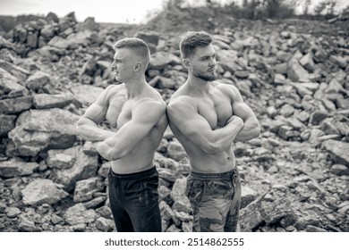 Two muscular men pose back-to-back in a rocky outdoor setting. Two men with well-defined muscles stand confidently back-to-back amid rocky terrain - Powered by Shutterstock