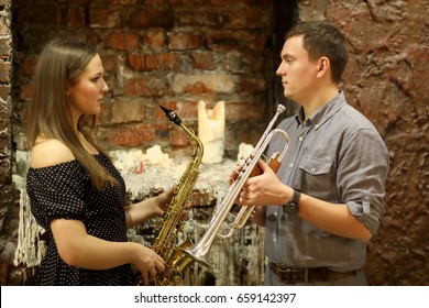 Two musicians with wind instruments in their hands look at each other in front a brick wall with a candle and a wax coating