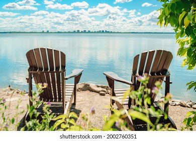 Two Muskoka chairs by the water on home terrace with calm view of lake in Canada. Summer cottage vacation lifestyle.
