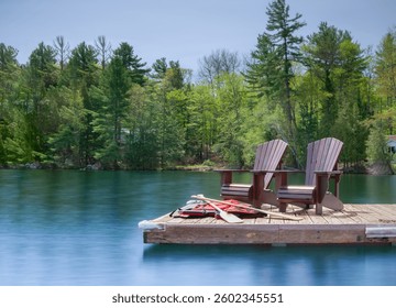Two Muskoka chairs rest on a wooden dock overlooking a calm lake, captured in a long exposure. A white cottage stands behind them, with life jackets and canoe paddles scattered nearby. - Powered by Shutterstock - Get 15% off with code: PIKWIZARD15