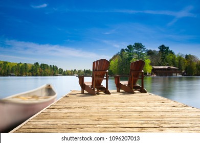 Two Muskoka chairs sitting on a wood dock facing a lake. Across the calm water is a brown cottage nestled among green trees. A canoe is tied to the dock.