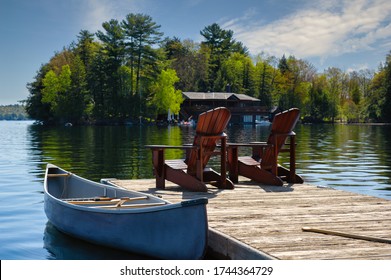 Two Muskoka chairs sitting on a wood dock facing a lake. Across the calm water is a brown cottage nestled among green trees. A canoe is tied to the dock.