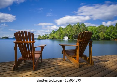 Two Muskoka chairs sitting on a wood dock facing a calm lake. Across the water is a white cottage nestled among green trees. There is a boat dock on the water in front of the cottage.
