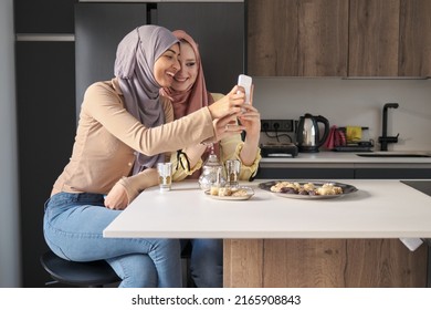 Two Muslim Women Laughing And Using The Smartphone While Having Tea And Arabic Sweets At The Kitchen Table.