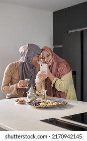 Two Muslim Women Laughing And Using The Smartphone While Having Tea And Arabic Sweets At The Kitchen Table.