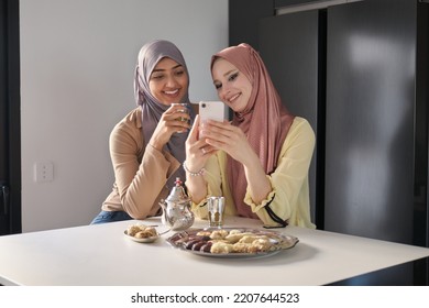 Two Muslim Women Laughing And Using The Smartphone While Having Tea And Arabic Sweets At The Kitchen Table.