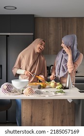 Two Muslim Women Laughing While Slicing, Chopping And Peeling Vegetables At Kitchen To Prepare A Salad.