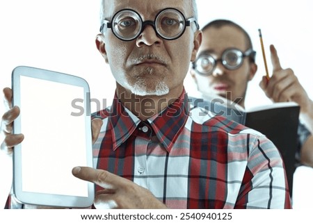 Two nerdy scientists with thick glasses are holding and pointing at a digital tablet with a blank screen, while one of them is taking notes on a book