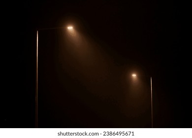 Two night lampposts shines with faint mysterious yellow light through evening fog. Streetlights shine at quiet city night, magic atmospheric light in mystical darkness