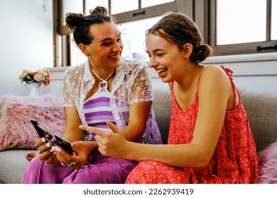 Two non binary friends sitting on couch looking at instant photos and laughing - Powered by Shutterstock