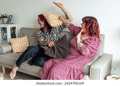 Two non-binary people are having fun hitting each other with pillows, sitting on their sofa in the living room, celebrating body positivity, gender equality and diversity
