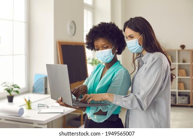 Two Office Coworkers In Medical Face Masks Using Laptop Together. Young Business Women Discussing New Project. Returning To Work After Covid-19 Lockdown. Safety Measures During Coronavirus Pandemic