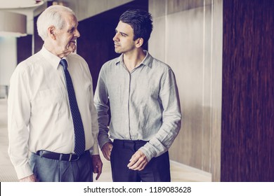 Two Office Employee Walking Along Office Space. Junior Employee Listening To Senior Mentor In White Shirt And Tie. Business Meeting And Coaching Concept