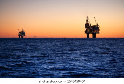 Two Oil Rigs In Silhouette During Sunrise In The Norwegian Oil Sector Of The North Sea