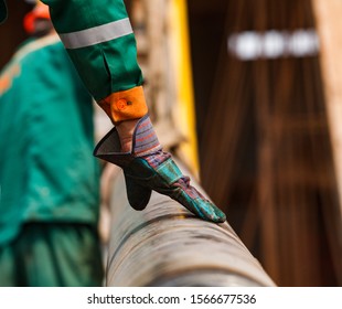 Two Oil Workers In Green And Orange Work Wear And Suede Gloves On Oil Deposit With Rusted Drilling Pipe. Close-up Of One Worker's Hand. Kazakhstan.