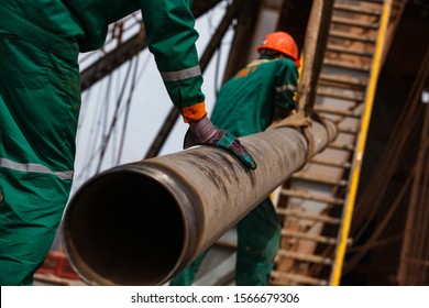 Two Oil Workers In Green And Orange Work Wear And Work Gloves With Rusted Drilling Pipe. Oil Deposit Zhaikmunai, Uralsk Region,  Kazakhstan.