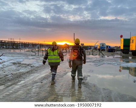 Two oil workers in work clothes walk through the site along a muddy road to an oil rig in the background of the sunset. Heavy male work of drillers.