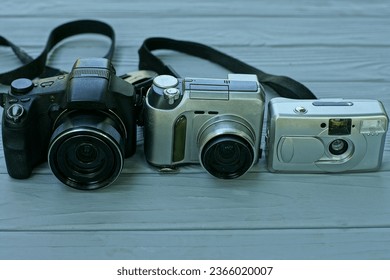  two old electronic black and white compact color ones with plastic and glass lenses, digital on batteries and one historical white film camera stand on a wooden gray table