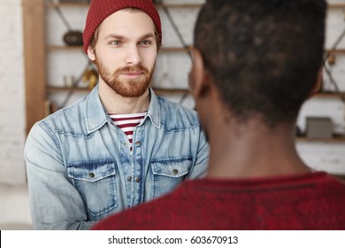 Two old friends of different races having nice conversation, sitting in front of each other at modern cafe, talking and sharing news. Stylish young Caucasian and African men chatting indoors - Powered by Shutterstock