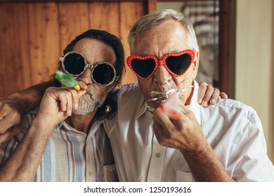 Two Old Friends With Party Blowers. Senior Men Wearing Funny Sunglasses Blowing Party Whistles Indoors.