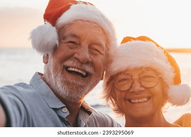 Two old happy seniors wearing christmas hats at the beach taking a selfie of them smiling and having fun with the sunset at the background at evening. Cute couple of old persons looking at the camera  - Powered by Shutterstock