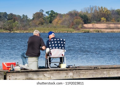 Two Old Men Fishing From A Dock. Early Fall Scene Of Elderly Friends Sitting On A Pier And Fishing