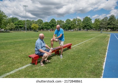 Two Old Men Are Having A Conversation Outdoors On A Sports Field.  One Man Is Sitting Whilst The Other Is Standing With One Leg Up On The Bench.  This Is A Married, Gay Couple.