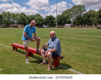 Two Old Men Are Having A Conversation Outdoors On A Sports Field. One Man Is Sitting Whilst The Other Is Standing With One Leg Up On The Bench. This Is A Married, Gay Couple.