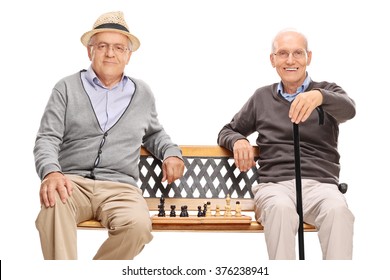 Two Old Men Posing Sitting On A Wooden Bench With A Chessboard Between Them Isolated On White Background