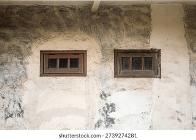 Two old, small rectangular wooden framed windows are set into a rough, textured concrete wall showing signs of age and weathering with large patches of discolored plaster