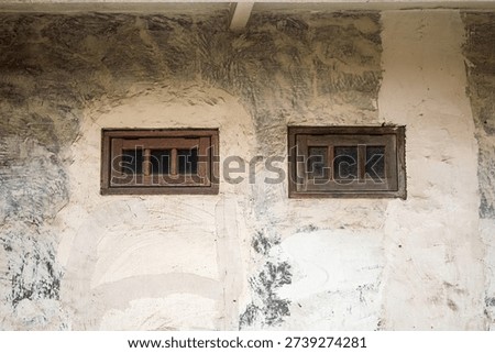Two old, small rectangular wooden framed windows are set into a rough, textured concrete wall showing signs of age and weathering with large patches of discolored plaster