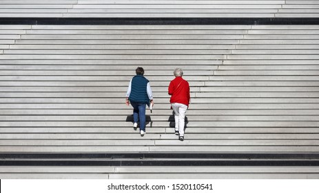Two Old Women Dressed In Blue And Red Climbing Up Big Stairs.