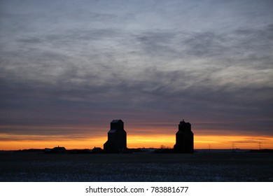 Two old wooden grain elevators silhouetted under a sunset sky in a countryside winter landscape