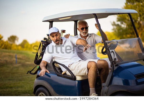 Two older\
friends are riding in a golf\
cart.