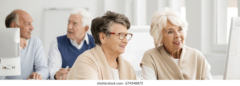 Two Older Women Sitting In Classroom With Friends Using Computer  