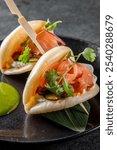 Two open-faced bao buns with salmon slices and herbs on a dark slate, accompanied by a green sauce. Perfect for a gourmet food concept or Asian cuisine theme.