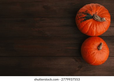 Two orange pumpkins are placed on a dark wooden table. The larger pumpkin is on top of the smaller one. This scene shows the colors of autumn and the harvest period.