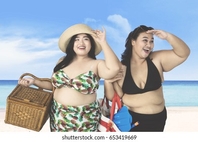 Two Overweight Women Looking Something On The Beach While Carrying Picnic Basket And Beach Items