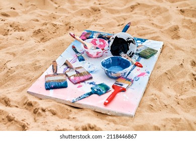 Two Painted Canvas In Blue And Pink Colors With Palettes Of Paints Lying On Sandy Beach, Artist's Palette At Outdoor Art Festival. White Drawing Canvas With Paint In Bowls, Art Performance Concept