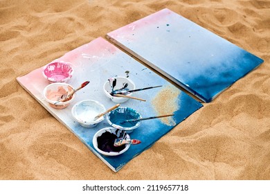 Two Painted Canvas In Blue And Pink Colors With Palettes Of Paints Lying On Sandy Beach, Artist's Palette At Outdoor Art Festival. White Drawing Canvas With Paint In Bowls, Art Performance Concept