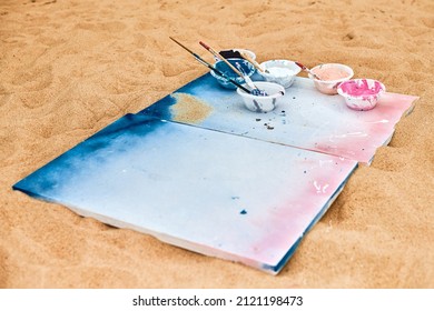 Two Painted Canvas In Blue And Pink Colors With Palettes Of Paints Lying On Sandy Beach, Artist's Palette At Outdoor Art Festival. White Drawing Canvas With Paint In Bowls, Art Performance Concept