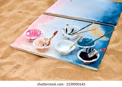 Two Painted Canvas In Blue And Pink Colors With Palettes Of Paints Lying On Sandy Beach, Artist's Palette At Outdoor Art Festival. White Drawing Canvas With Paint In Bowls, Art Performance Concept