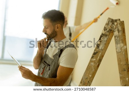 Two painters focus on interior wall renovation. One assesses plans with a thoughtful expression while the other applies paint from a roller on a ladder.