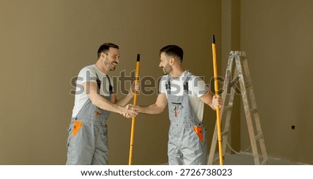 Two painters are shaking hands and smiling, holding long paint rollers in a freshly painted room. A ladder is visible nearby, indicating ongoing renovation work in a warm-colored space.
