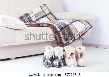 Two pair of natural woollen slippers on wooden floor and blanket on the couch