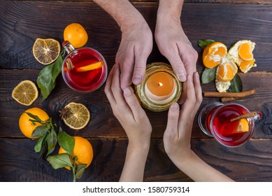 Two Pairs Of Hands, Male And Female, Holding A Burning Scented Candle Together. Standing Next To A Glass Cups With Mulled Wine. Cozy Flatlay On A Wooden Background In Dark Christmas Style.