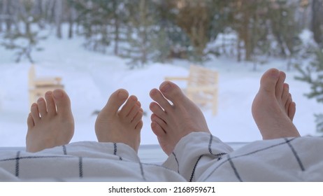 Two Pairs Of Legs, Male And Female Bare Feet Under A Blanket. A Woman With A Man Under A Blanket In Front Of A Window With A Winter Landscape. Couple In Love Under A Blanket Together.